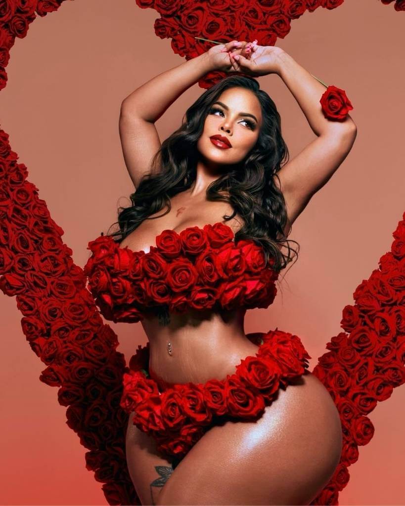 Portrait of Gracie Bon with long dark hair, dressed in a white top and denim jacket, smiling softly at the camera. (1) Gracie Bon is wearing red roses on her body